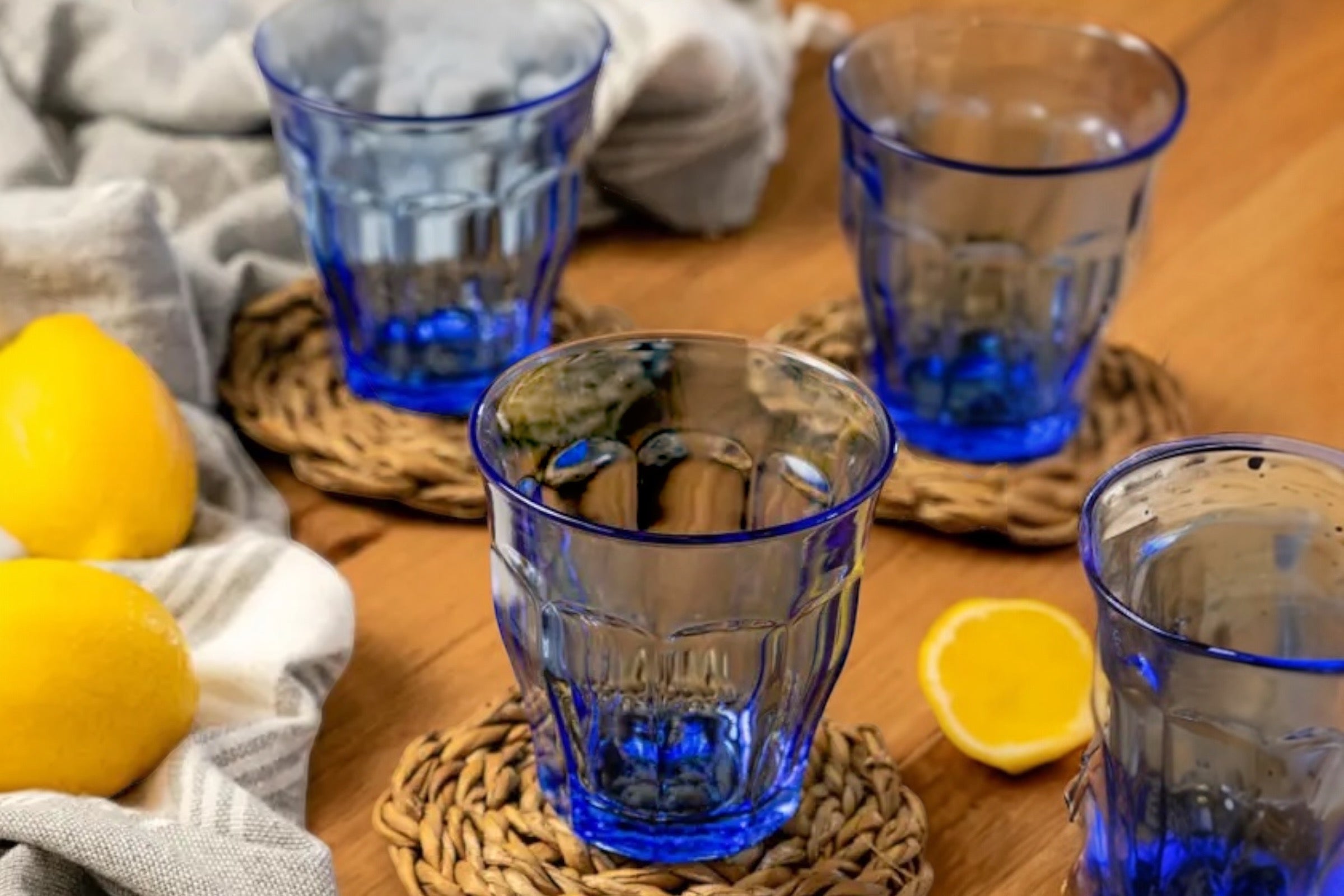 Blue glass tumblers on woven coasters with lemons on a wooden table.