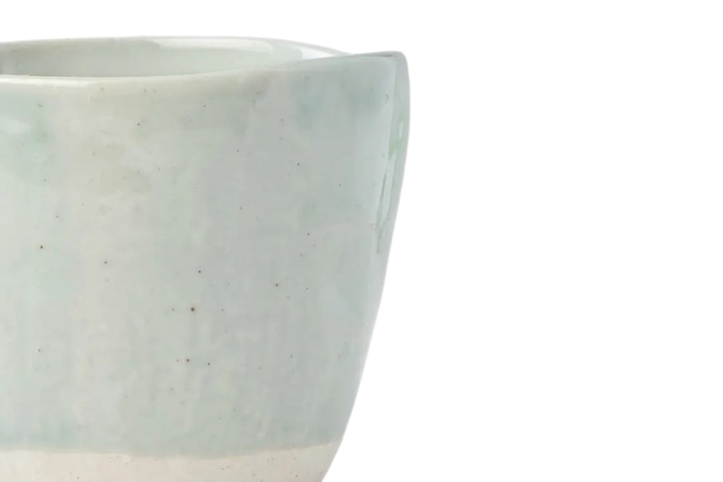 A close up of a hand-finished ceramic mug with a softly irregular form and pale tomei blue glaze set against a white background.
