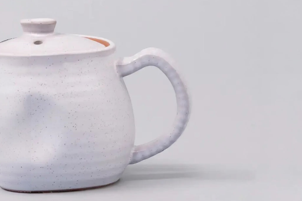 A cut off photo of a white speckled japanese teapot with a ridge design, set against a white background.