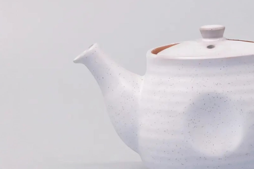 A cut off photo of a white speckled japanese teapot with a ridge design, set against a white background.
