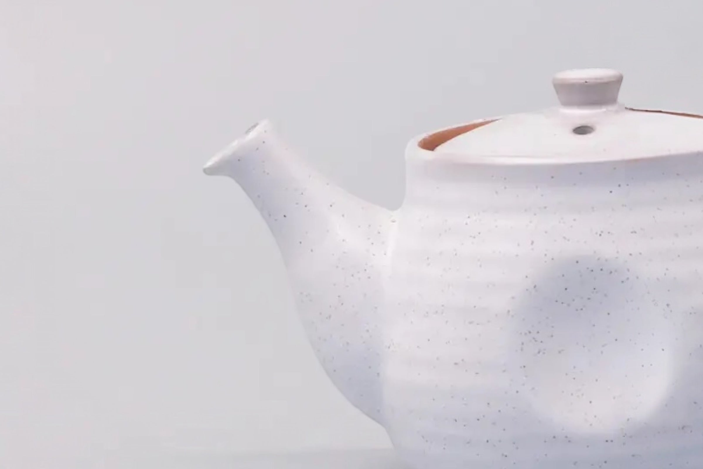 A cut off photo of a white speckled japanese teapot with a ridge design, set against a white background.